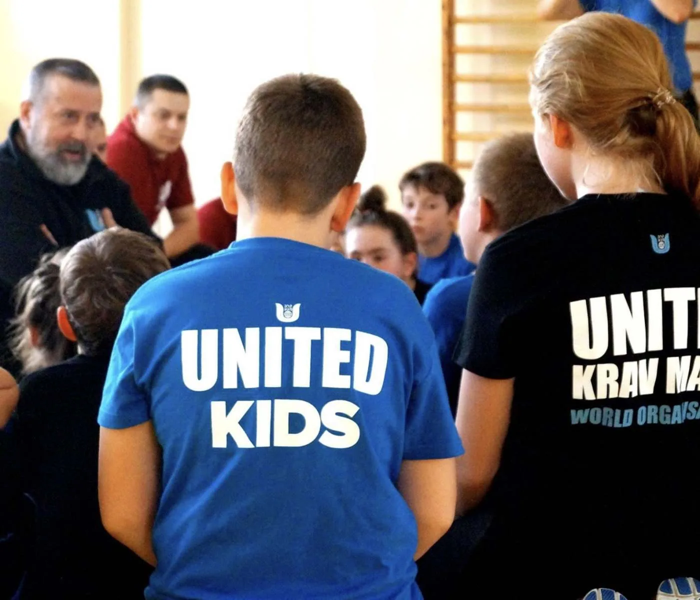 Children gather in a gym with mentors; two wear “United Kids” and “United Krav Maga” shirts, creating a friendly, team-focused mood.