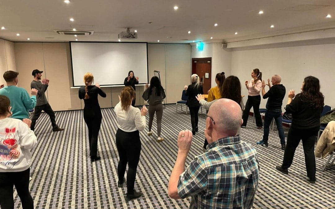 Participants follow an instructor’s movements during an upbeat group activity in a hotel conference room, with chairs and a projector screen.